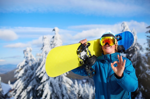 Snowboarder sorridente posando carregando snowboard nos ombros na estância de esqui perto da floresta antes da sessão de freeride Rider mostrando sinal de OK usando óculos polarizados Equipamento de snowboard moderno