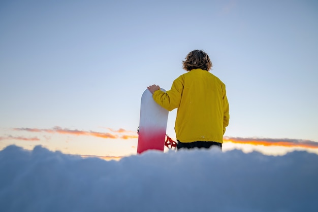 Snowboarder sosteniendo una tabla de snowboard en la cima de la montaña y mirando reflejándose en la distancia