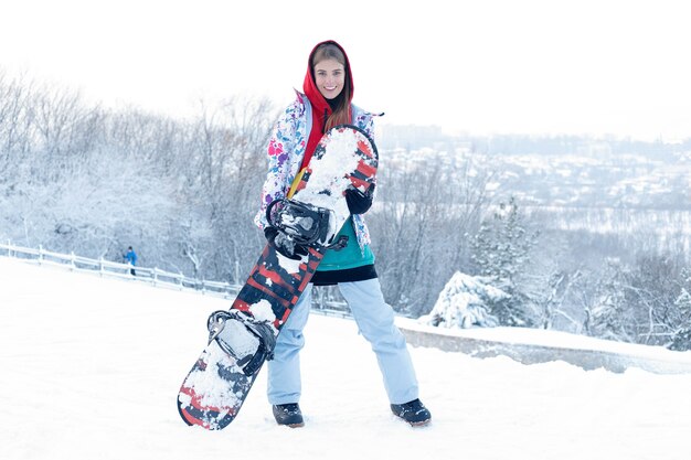 Snowboardkonzept des Frauenwinters im Freien. Junge Frau mit Snowboard auf den Schultern, sie schaut weg und lächelt, Kopienraum, Nahaufnahme