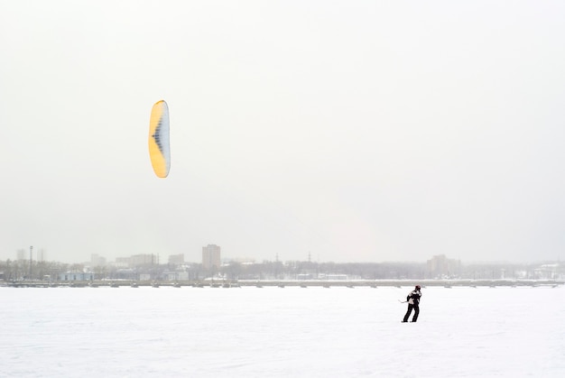 Snowkiter fährt bei Schneefall auf dem Eis eines zugefrorenen Sees vor dem Hintergrund der Stadt