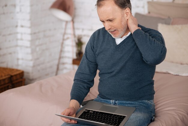 So schlimmer Schmerz. Alter Mann, der Schmerzen im Nackenbereich fühlt, während er auf dem Bett sitzt und Laptop verwendet.