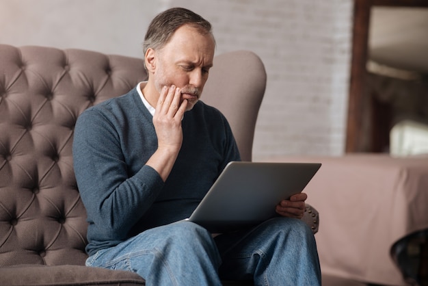 So schmerzhaft. Porträt des älteren gutaussehenden Mannes mit starken Zahnschmerzen beim Sitzen auf dem Sofa und beim Verwenden des Laptops zu Hause.