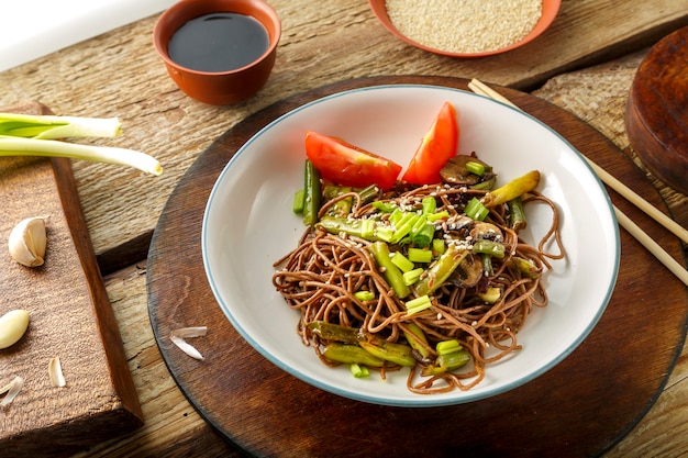 Soba mit Pilzen, grünen Bohnen und Sesam in einem Teller auf einem Ständer auf einem Holztisch neben Stäbchen und Sojasauce und Knoblauch auf Ständern. Horizontales Foto