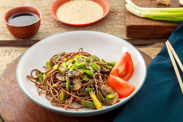 Soba mit pilzen, grünen bohnen und sesam in einem teller auf einem ständer auf einem holztisch neben stöcken und sojasauce und sesam. horizontales foto.