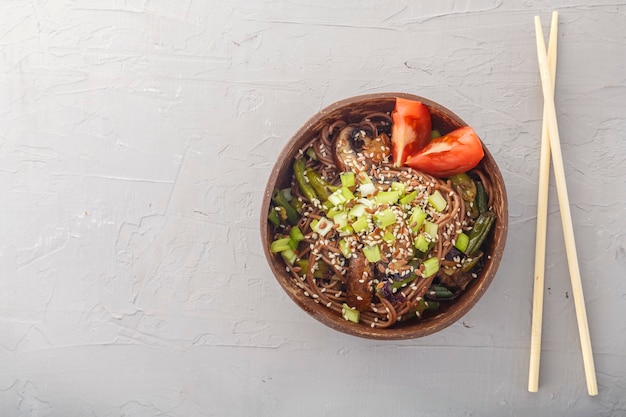 Soba con setas y semillas de sésamo en un cuenco de cáscaras de coco sobre un fondo de hormigón cerca de palos. Foto horizontal