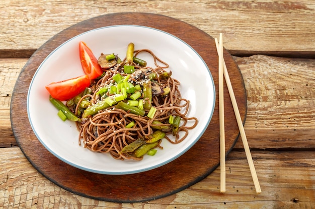 Soba con setas y semillas de sésamo en un plato sobre un soporte de madera sobre la mesa junto a los palillos. Foto horizontal