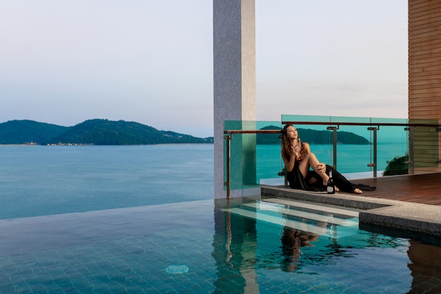 Soberba jovem garota em um vestido preto longo senta-se com uma taça de champanhe perto da piscina de beiral infinito, excelente vista para o mar