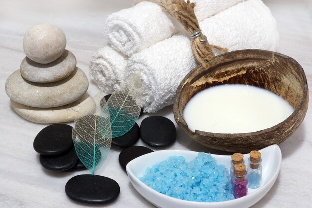 Sobre a bancada de mármore branco encontra-se um conjunto para procedimentos de Spa com leite de coco, leite rejuvenescedor, pedras e sal de banho azul.