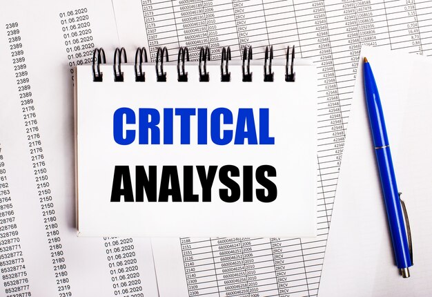 Sobre a mesa estão gráficos e relatórios, nos quais estão uma caneta azul e um caderno com a palavra ANÁLISE CRÍTICA