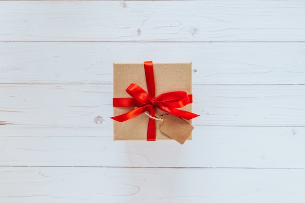 Sobre la caja de regalo marrón con la etiqueta en fondo de la tarjeta de madera. Caja de regalo con cinta roja sobre fondo blanco de madera con espacio.