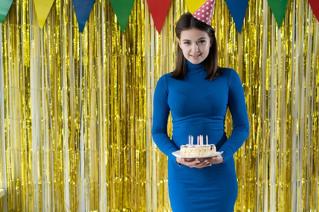 Sobre un fondo amarillo, una mujer con un vestido azul está de pie sosteniendo un pastel con velas en su sombrero de fiesta en la cabeza mirando a la cámara Baner