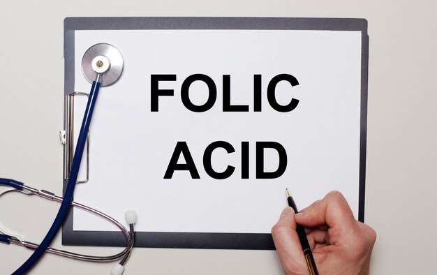 Sobre un fondo claro, un estetoscopio y una hoja de papel, en la que un hombre escribe ÁCIDO FÓLICO. Concepto medico
