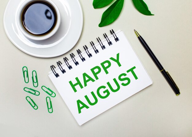 Sobre un fondo gris claro, una taza de café blanca, clips verdes y una hoja verde de una planta, así como un bolígrafo y un cuaderno con las palabras FELIZ AGOSTO.