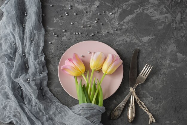 sobre un fondo gris en un plato rosado hay tulipanes rosados, perlas grises y cubiertos - cuchillo y tenedor