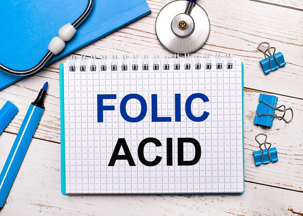 Sobre un fondo de madera clara, un estetoscopio, un bloc de notas azul, clips de papel azules, un marcador azul y una hoja de papel con el texto ÁCIDO FÓLICO Concepto médico