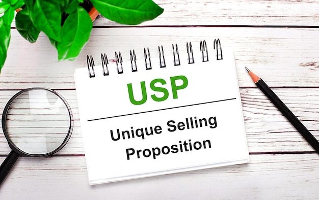 Sobre un fondo de madera clara, una lupa, un lápiz, una planta verde y un cuaderno blanco con texto USP Unique Selling Proposition Business concept