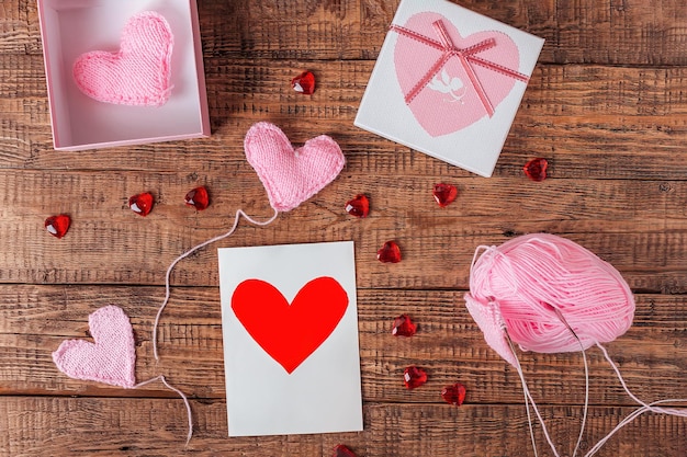 Sobre un fondo de madera, corazones rosas de punto, herramientas de tejer, una caja de recuerdos y una tarjeta de felicitación