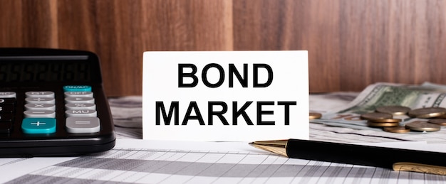 Sobre un fondo de madera se encuentra un bolígrafo con una calculadora y una tarjeta blanca con las palabras BOND MARKET