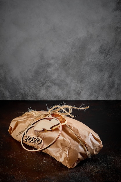 Sobre un fondo de textura oscura, un paquete de regalo hecho de papel Kraft atado con una cuerda con el emblema del ratón símbolo de año nuevo hecho de madera contrachapada