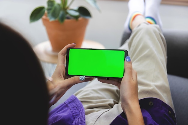 Sobre el hombro Chica usando una computadora móvil con teléfono inteligente en el sofá en la sala de estar con pantalla verde en blanco Concepto de trabajo desde casa