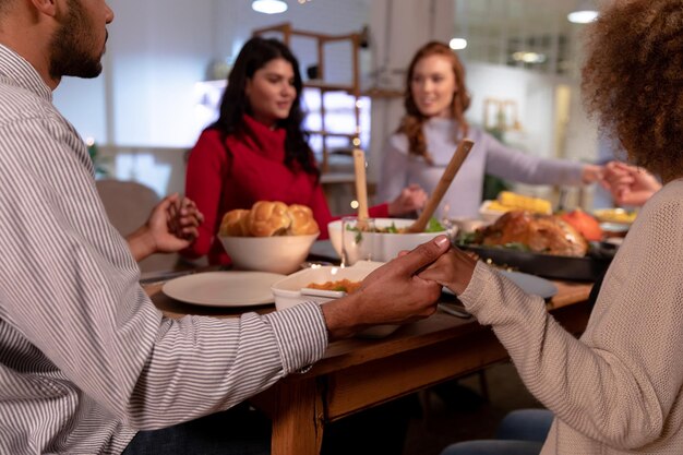 Sobre el hombro vista de un grupo de adultos jóvenes multiétnicos hombres y mujeres amigos sentados alrededor de una mesa de la mano diciendo la gracia antes de comer la cena de Acción de Gracias en casa juntos