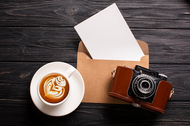 Un sobre con un lugar para una inscripción, una hermosa cámara y una taza de café con espuma sobre un fondo de madera negra
