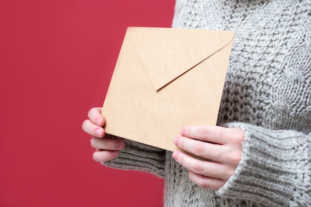 Sobre marrón de papel Kraft en manos de mujeres sobre un fondo rojo, primer plano. Una carta a Santa, concepto de lista de deseos. La niña sostiene una carta. Fondo de año nuevo.