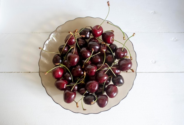 Sobre una mesa blanca hay un plato de vidrio con cerezas oscuras cerezas negras en un plato sobre una mesa de madera Cosecha de cerezas útiles bayas de verano