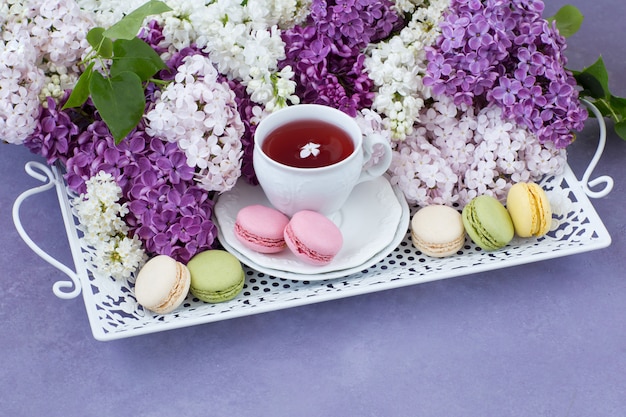 Sobre una mesa de color lila sobre una vieja bandeja blanca, una taza de té, macarrones y un ramo de lilas.