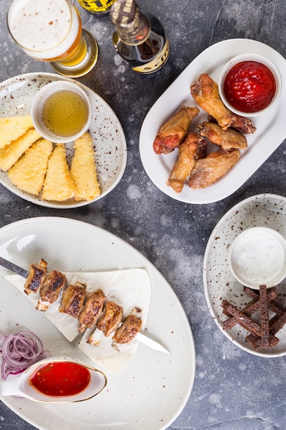 Sobre la mesa hay jugosos shish kebab y muslos de pollo quemados con salsa de tomate picante, crutones y queso frito en pan rallado y un vaso de cerveza lleno.