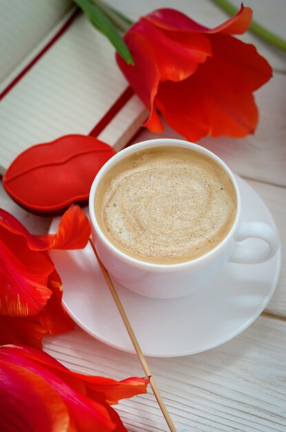 Sobre una mesa de madera blanca hay un cuaderno rojo con tulipanes, hay una taza de café y pan de jengibre en forma de labios escarlata.