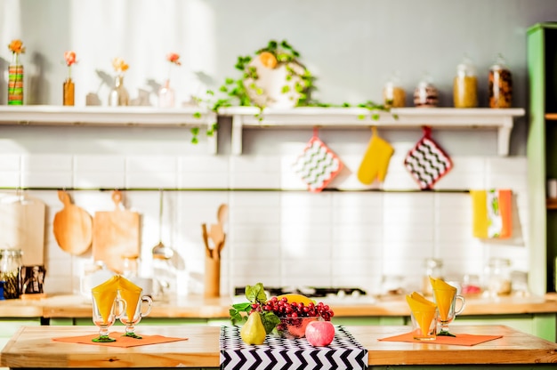 Sobre una mesa de madera hay un cuenco de fruta, vasos de cristal con servilletas. En el fondo hay un interior de cocina decorado con estilo. Foto horizontal