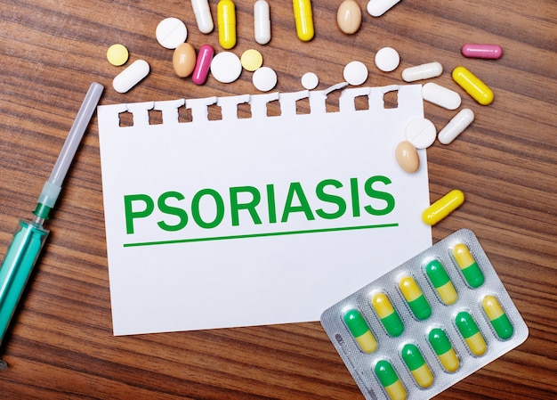 Sobre una mesa de madera, una jeringa, pastillas y una hoja de papel con la inscripción PSORIASIS. Concepto medico