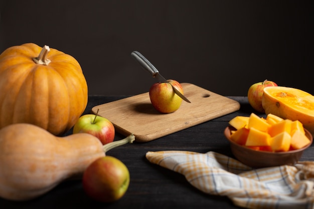 Sobre una mesa de madera negra se encuentran calabazas de varios tamaños y formas, manzanas maduras, un paño de cocina y un tazón de calabaza en rodajas.