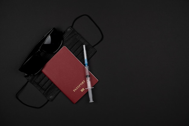 Sobre una mesa negra, un pasaporte, gafas negras, una máscara médica negra y una jeringa. Concepto de viaje en cuarentena