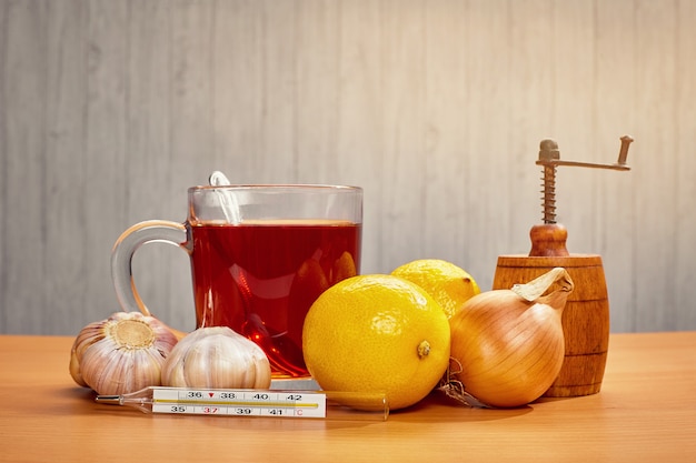 Sobre la mesa una taza de té con limón, cebolla, ajo y un termómetro.