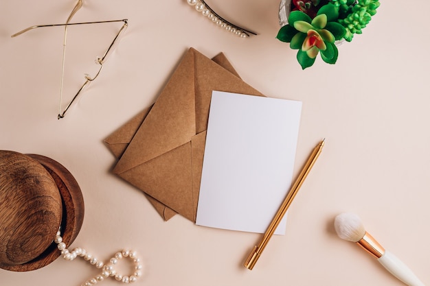 Sobre de papel artesanal con maqueta de nota de papel en blanco blanco con pinceles de maquillaje, gafas y perlas sobre fondo beige. Vista plana endecha, superior. Invitación, paquete y carta