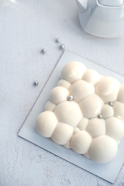 Foto sobre un plato blanco, una tetera y una tarta de mousse con relleno de fresa con una capa blanca aterciopelada en forma de burbujas