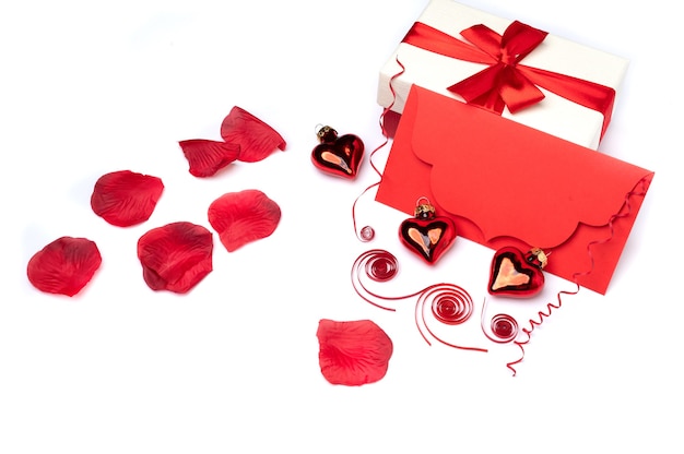 Sobre rojo de felicitación sobre una superficie blanca decorada con dos corazones rojos de cristal al estilo del Día de San Valentín y pétalos de rosa hay un lugar para el texto