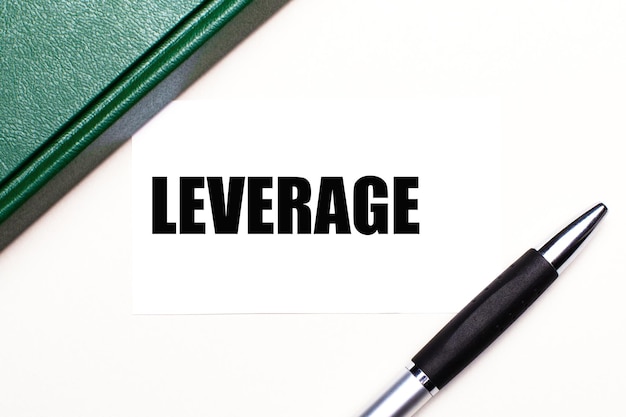 Sobre um fundo cinza claro, encontra-se uma caneta, um caderno verde e um cartão branco com o texto leverage business concept