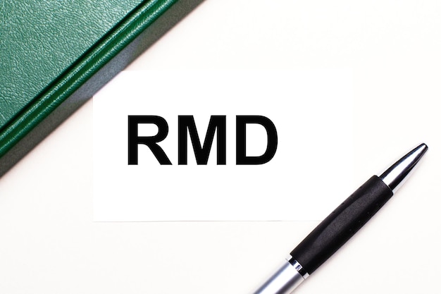 Foto sobre um fundo cinza claro, encontra-se uma caneta, um caderno verde e um cartão branco com o texto rmd required minimum distributions business concept