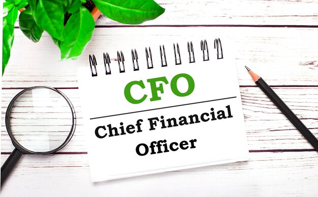 Sobre um fundo claro de madeira, uma lupa, um lápis, uma planta verde e um caderno branco com texto CFO Chief Financial Officer. Conceito de negócios