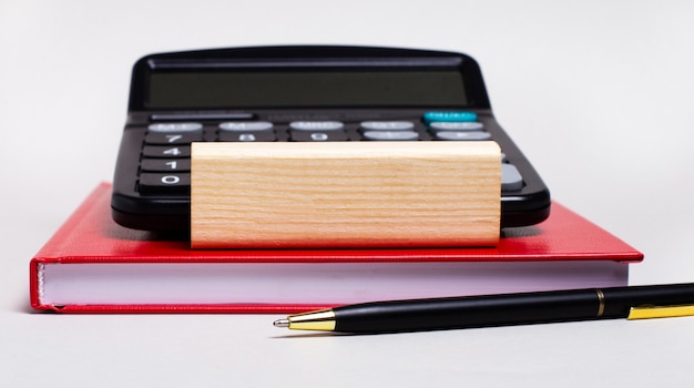 Sobre um fundo claro, um caderno cor de vinho, uma calculadora, uma caneta e um bloco de madeira com local para inserir texto.