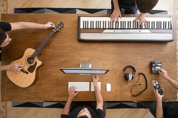 Sobre uma mesa de madeira de um estúdio de gravação, um teclado musical, um violão, uma mesa de som e um computador.