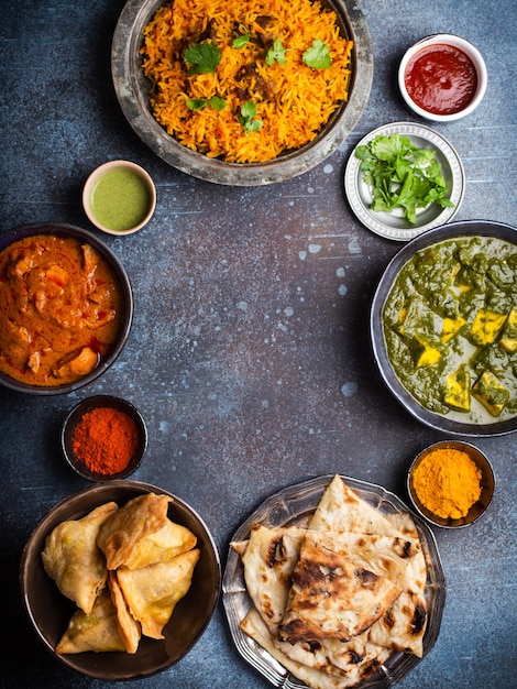 Sobrecarga de platos y aperitivos tradicionales indios: pollo al curry, pilaf, pan naan, samosas, paneer, chutney sobre fondo rústico. Mesa con elección de comida de la cocina india, espacio para texto