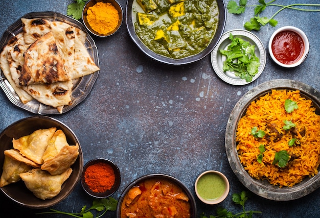 Sobrecarga de platos y aperitivos tradicionales indios: pollo al curry, pilaf, pan naan, samosas, paneer, chutney sobre fondo rústico. Mesa con elección de comida de la cocina india, espacio para texto