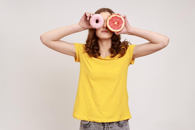 Sobremesa açucarada vs frutas. retrato de mulher de cabelos castanhos brincalhão em camiseta amarela cobrindo os olhos com toranja fresca e rosquinha doce, junk food. tiro de estúdio interior isolado em fundo cinza.