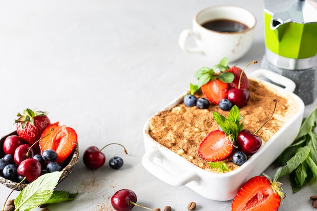 Sobremesa italiana clássica Tiramisu decorada com morangos, cerejas e hortelã em um fundo branco. Copie o espaço para o seu texto.