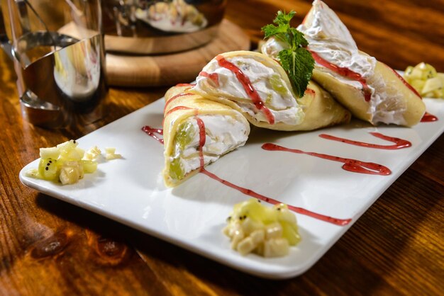 Sobremesa. Panquecas com creme e frutas. Decorado com calda e hortelã em uma mesa de madeira