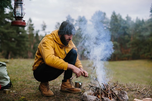 Sobrevivência na natureza Um homem barbudo acende uma fogueira perto de um abrigo improvisado feito de galhos de pinheiro